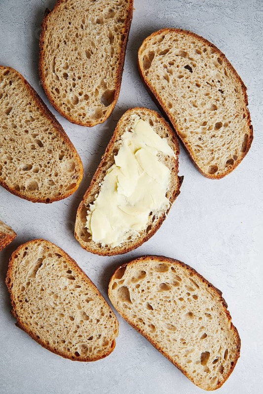 Top view of sliced fresh homemade sourdough bread with some slices generously spread with butter, set on a grey textured surface