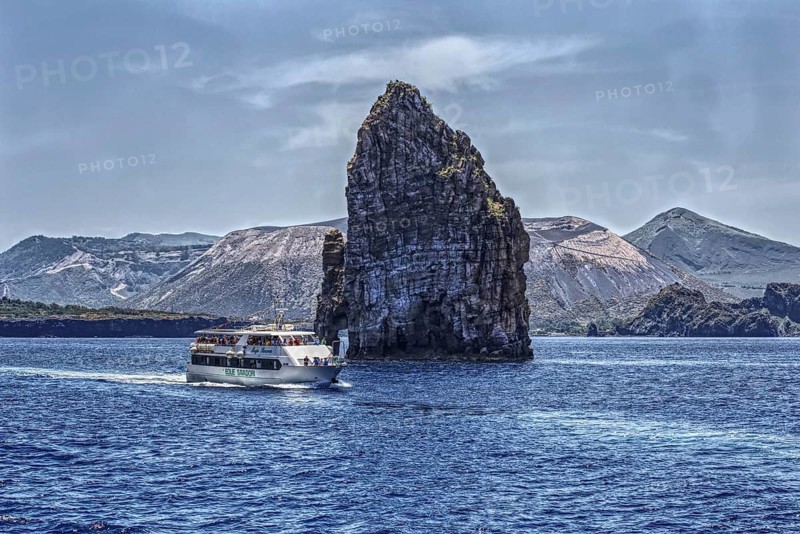 A large rock massif juts out of the blue sea, a ship sails past it, Lipari Aeolian Islands, Sicily Italy