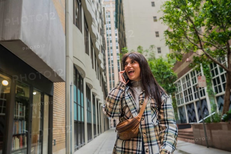 Young woman smiling and talking on her mobile phone while walking through a city alley lined with modern buildings, enjoying communication and casual urban exploration