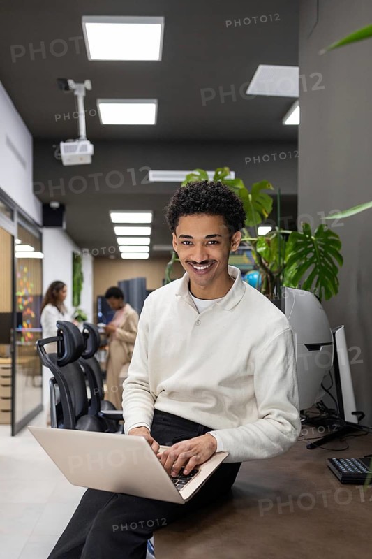 A cheerful professional sits on a desk in an open, modern office environment, working on a laptop. Colleagues in the background contribute to a collaborative atmosphere