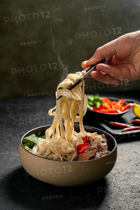 Cropped unrecognizable person enjoying Vietnamese Beef Pho Noodle Soup, with steam rising from the bowl as noodles are lifted with chopsticks