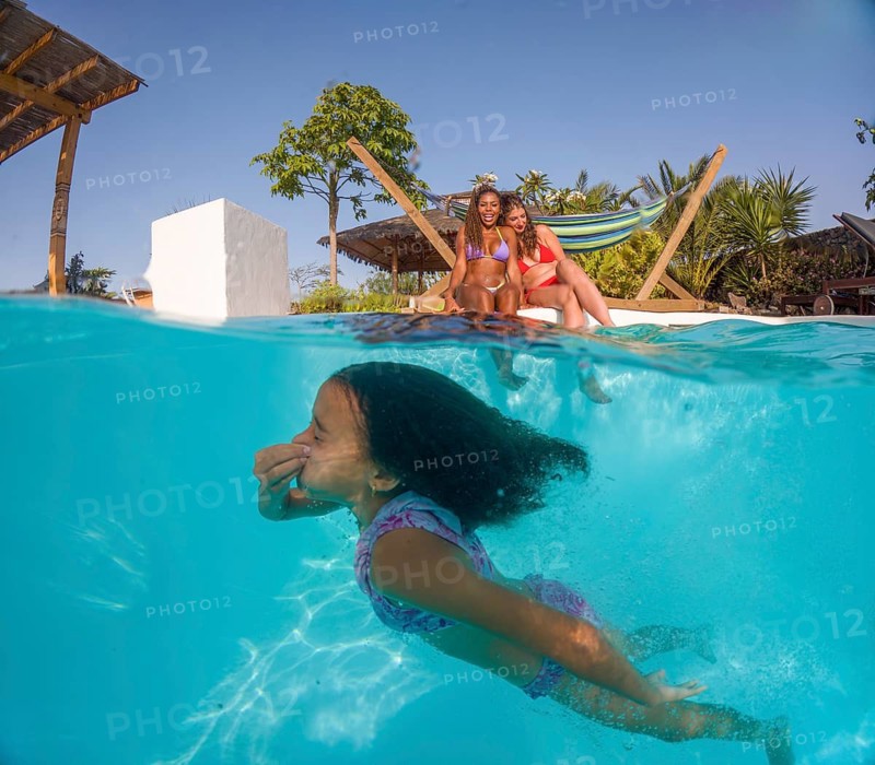 A loving lesbian family relaxes by the pool on a sunny day. A child joyfully swims underwater while two women smile brightly on the poolside, embracing love and diversity