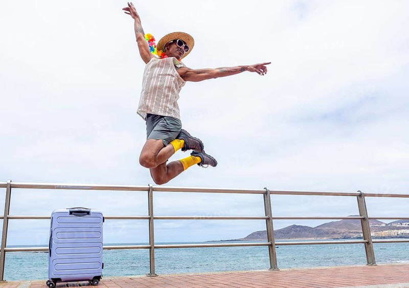 A joyful LGBTQIA+ man, with a suitcase, leaps in the air, excitedly preparing for travel and holidays by the ocean. Their energy embodies pride and freedom