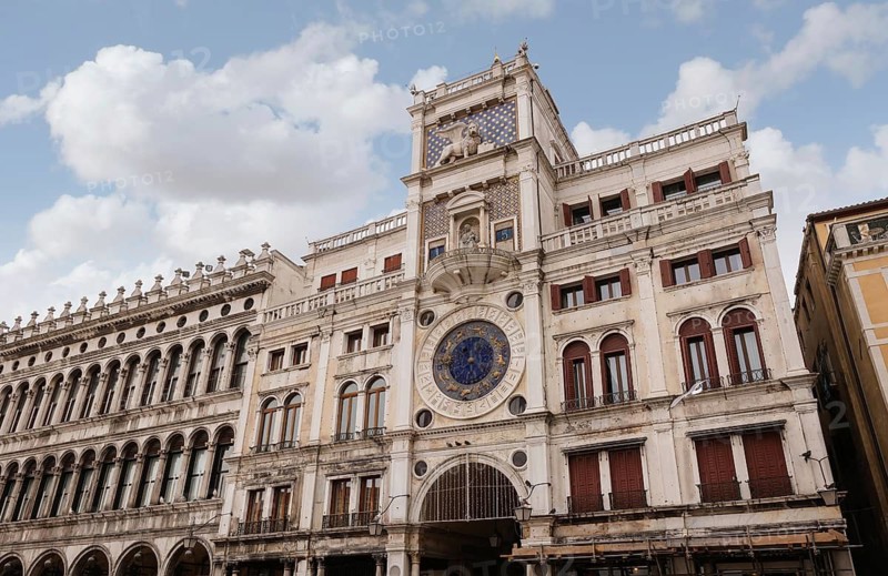 The historic clock tower in Venice, Italy, showcases intricate architectural details, with its ornate facade and large clock face against a backdrop of cloudy sky