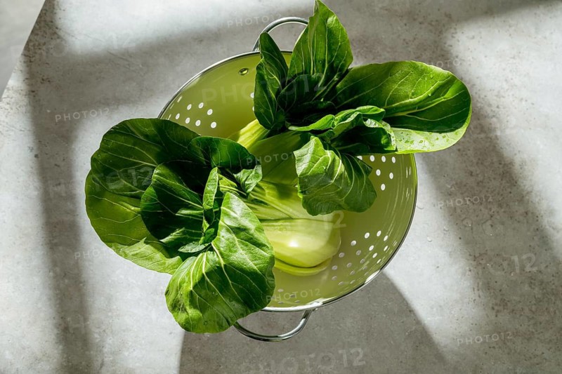 Fresh, vibrant bok choy rests in a colander, illuminated by soft natural light. The green leaves texture and shadows create an appealing and healthy visual