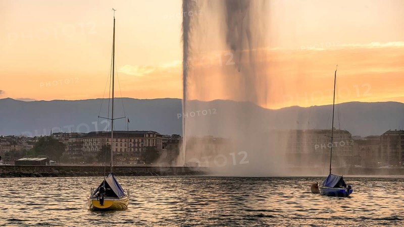 A breathtaking sunset view at Lake Geneva featuring the iconic Jet d'Eau fountain with colorful sailing boats on calm waters