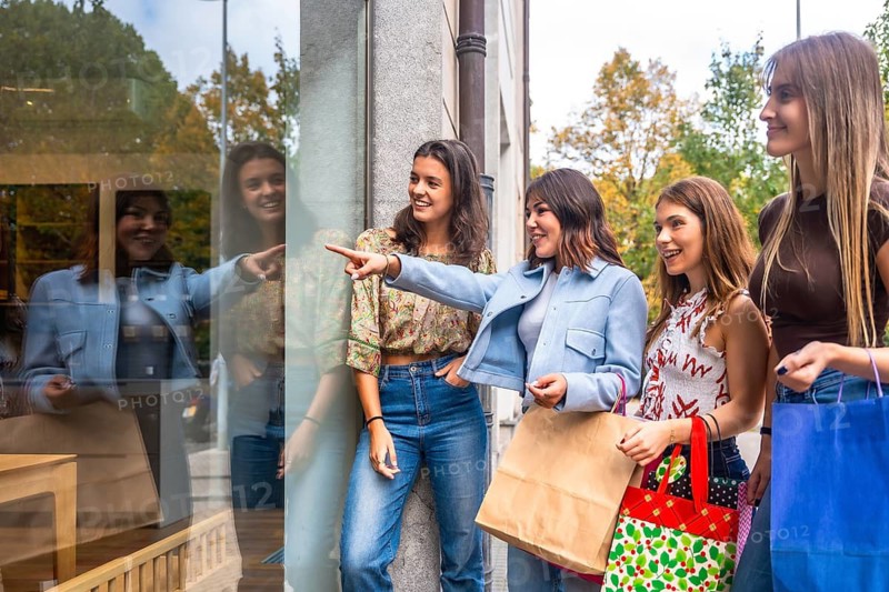 Group of young women friends happily window shopping, pointing and looking at items in a store display while carrying shopping bags on a casual day out in the city