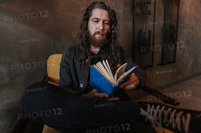 A man with long hair and a beard reads a blue book while sitting in a cozy chair of a cafe, creating a relaxed and intellectual atmosphere, perfect for study or leisure