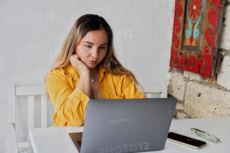 A young woman attentively using her laptop in a cozy cafe. With her hand resting on her chin, she is fully immersed in her work, surrounded by casual ambiance and decor