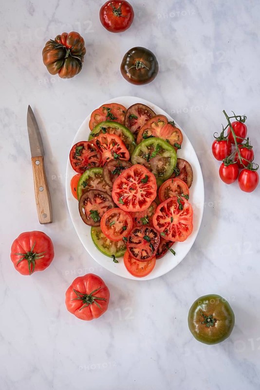 A colorful assortment of sliced heirloom tomatoes garnished with fresh herbs, arranged on a white plate. A knife and whole tomatoes are placed on a marble surface for a rustic presentation