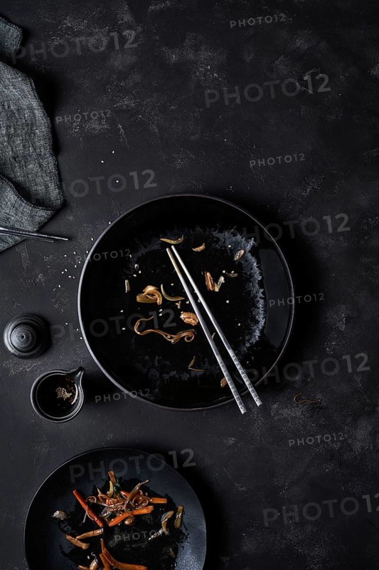 Overhead black empty bowl with pair of chopsticks and leftovers of Korean cuisine Japchae at table near full plate