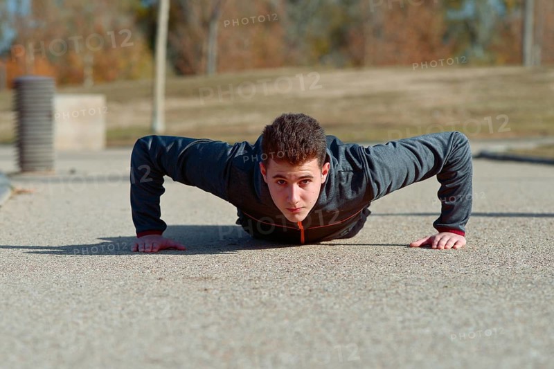 A young athlete performs push ups on a sunny day outdoors, exhibiting strength and focus. He is wearing a dark athletic top, with a park setting in the background