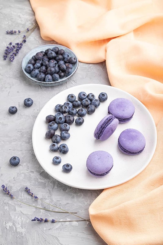 Purple macarons or macaroons cakes with blueberries on white ceramic plate on a gray concrete background and orange textile. Side view, close up