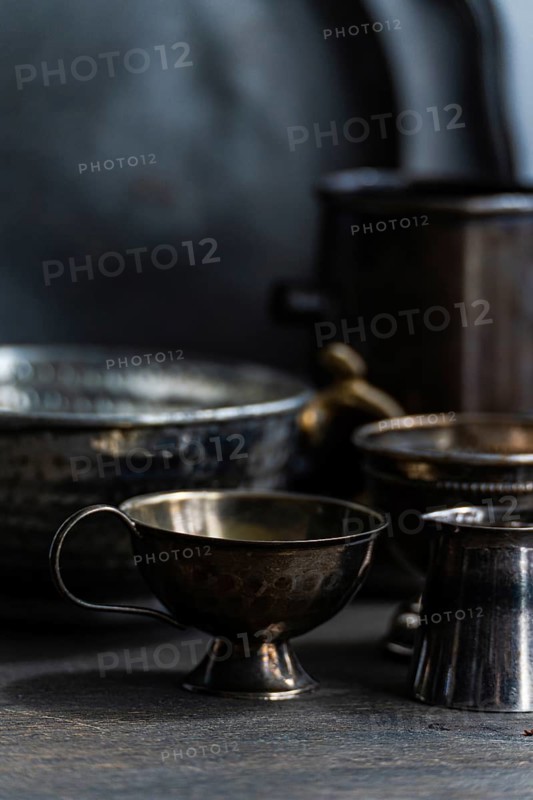 A collection of vintage metal cups with textured handles is displayed on a dark surface. The dim lighting highlights their rustic patina and intricate designs, evoking a sense of nostalgia