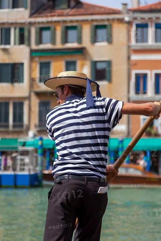 Gondolier in striped shirt steering boat along a Venetian canal in sunny weather, Venice, Italy