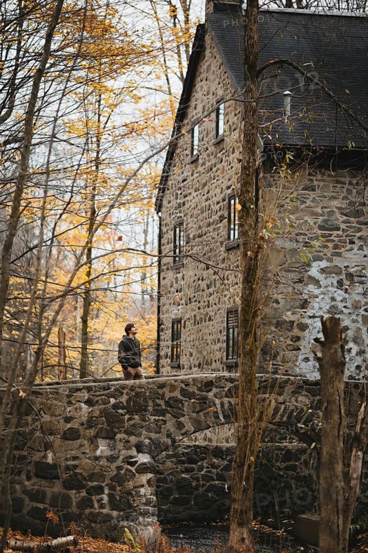 A man walks on a stone bridge near an old stone building in an autumnal forest in Quebec, Canada. Golden leaves create a picturesque landscape, perfect for seasonal reflection