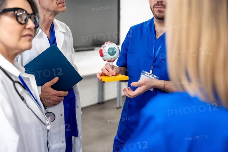 Healthcare team members, including doctors and nurses, collaborating and educating, standing together while holding and examining a human eye model during a discussion in a clinic or conference room