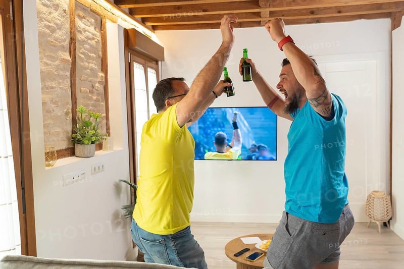 Friends reacting with excitement after a goal while watching a match on television. Spontaneous celebration, positive emotions and friendship indoors