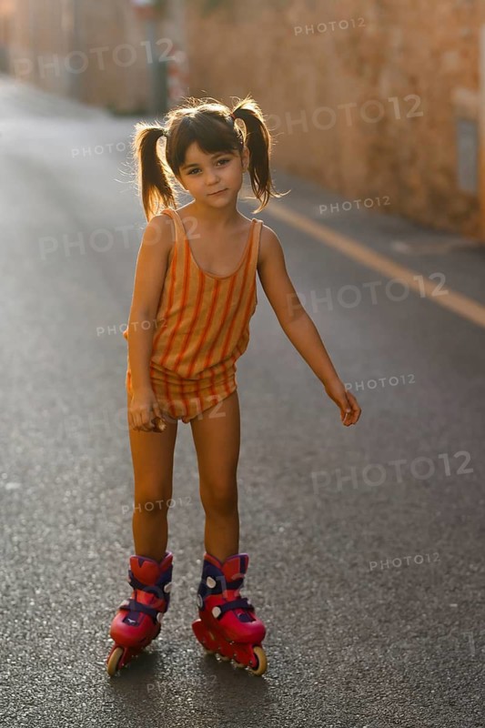 A young girl in a striped outfit enjoys rollerblading on a sunlit street in Santa Maria del Cami, Mallorca, capturing a carefree, playful moment