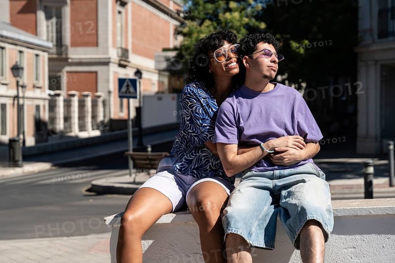 A joyful, multiethnic LGBTQ+ couple shares a tender moment while seated outdoors. The scene reflects love, diversity, and carefree happiness on a sunny day