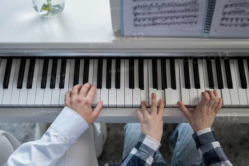 Top view of two pairs of hands playing a piano keyboard together, highlighting the essence of musical collaboration and learning, with sheet music visible in soft focus