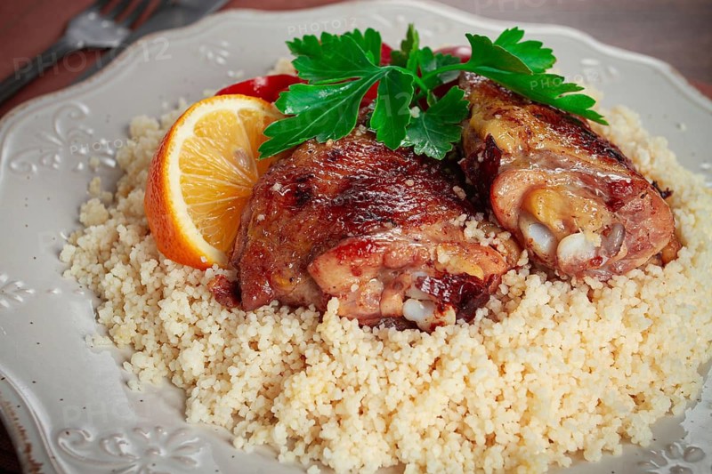 Couscous is served on a plate with pieces of chicken on top. Lemon slices and parsley decorate the dish