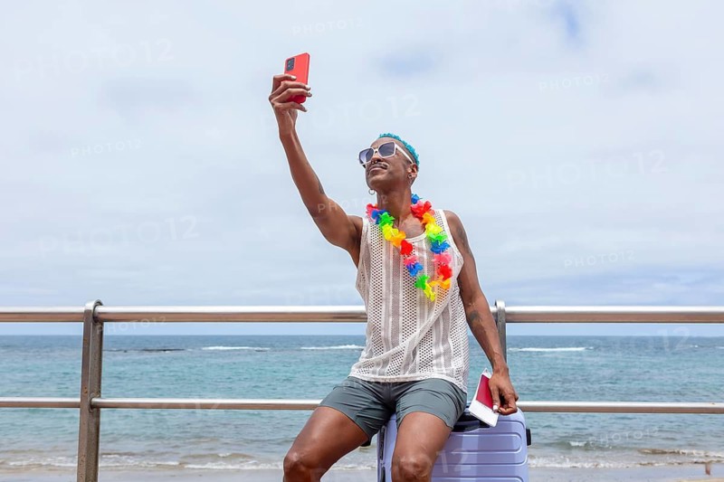 An LGBTQIA+ man sitting on a suitcase on the beach, adorned with a colorful flower necklace, takes a joyful selfie. Ready for a vacation, embracing freedom and pride