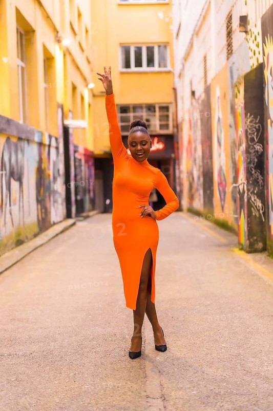 Fashionable urban style with a black African girl in an orange dress and black heels on a city street. Smiling and arm up