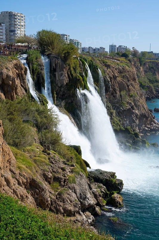 Dynamic waterfall flows into the sea surrounded by rocks and cityscape in the background, Lower Düden Waterfall, Düden River, Antalya, Turkey