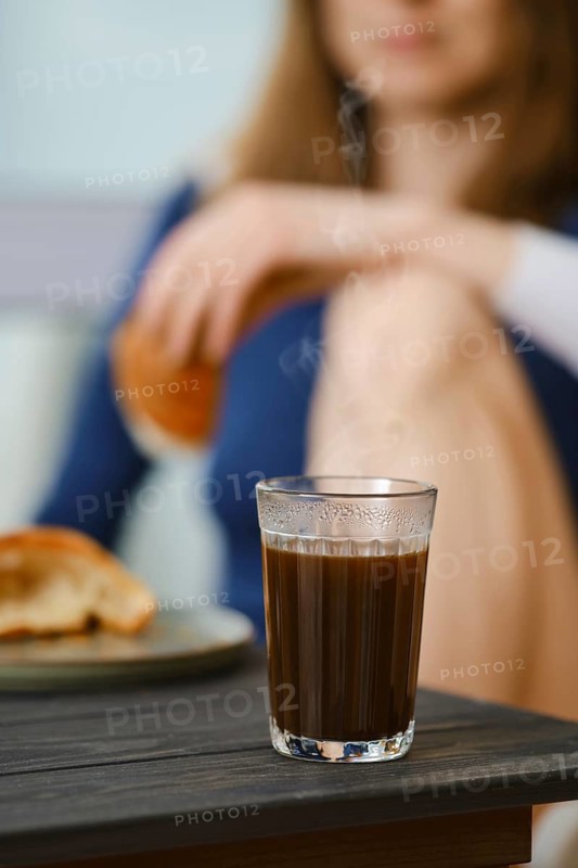 Coffee glass with unrecognizable blurred woman sitting in the kitchen with croissant in hand, focus on glass with coffee