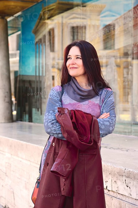 A woman stands confidently outdoors, wearing a colorful sweater and holding a maroon jacket. She is near a modern building with reflective glass under soft daylight