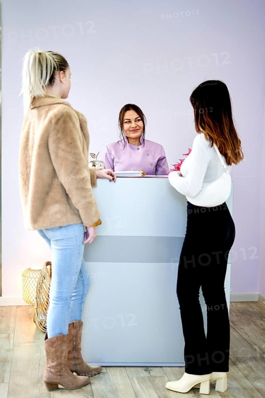 Two women engage in conversation with a professional at a beauty salon reception. The modern space features a welcoming atmosphere, emphasizing personal care and beauty services