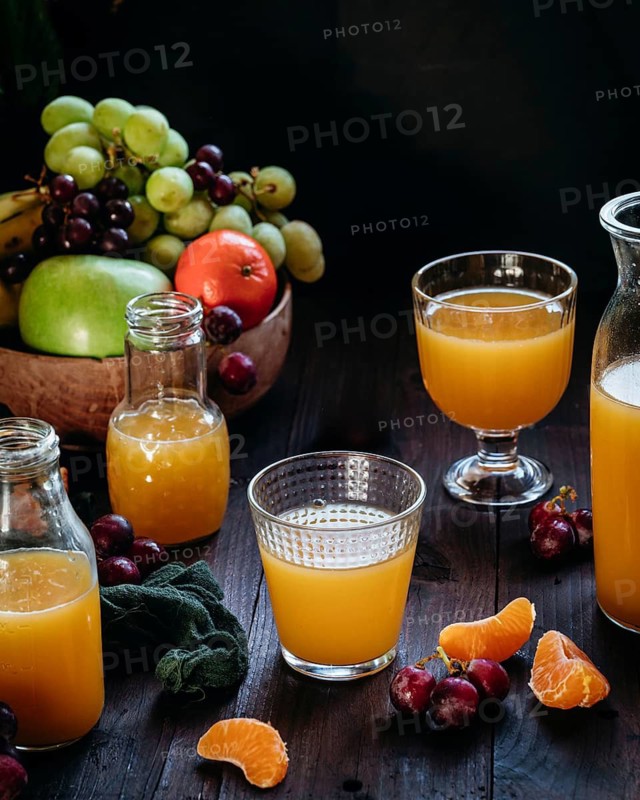 Assorted glasses and bottles of fresh orange juice surrounded by vibrant fruits, including grapes and oranges, on a rustic wooden table, creating a refreshing visual