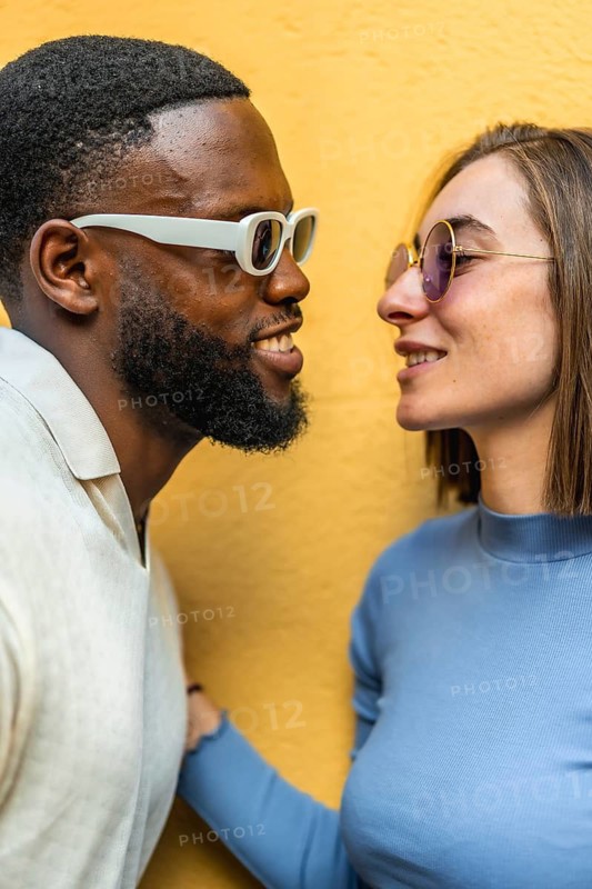 Vertical close-up photo of a happy cool modern multi-ethnic couple with sunglasses about to kiss against yellow background