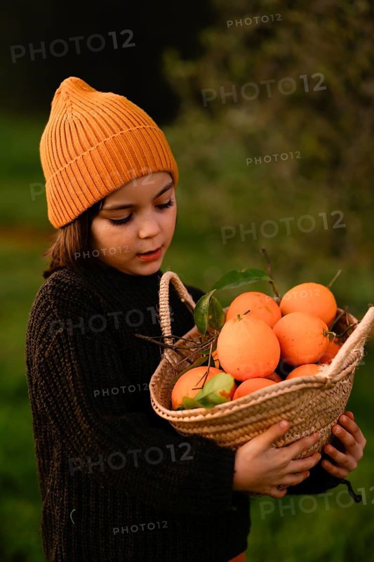 A young child in a cozy orange beanie and dark sweater holds a woven basket filled with fresh oranges. The green, natural background enhances the vibrant colors