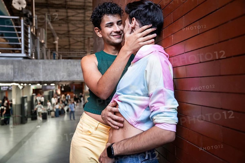 A gay couple shares a tender moment in a train station, showcasing love and diversity. The vibrant colors and urban backdrop emphasize a modern romantic scene