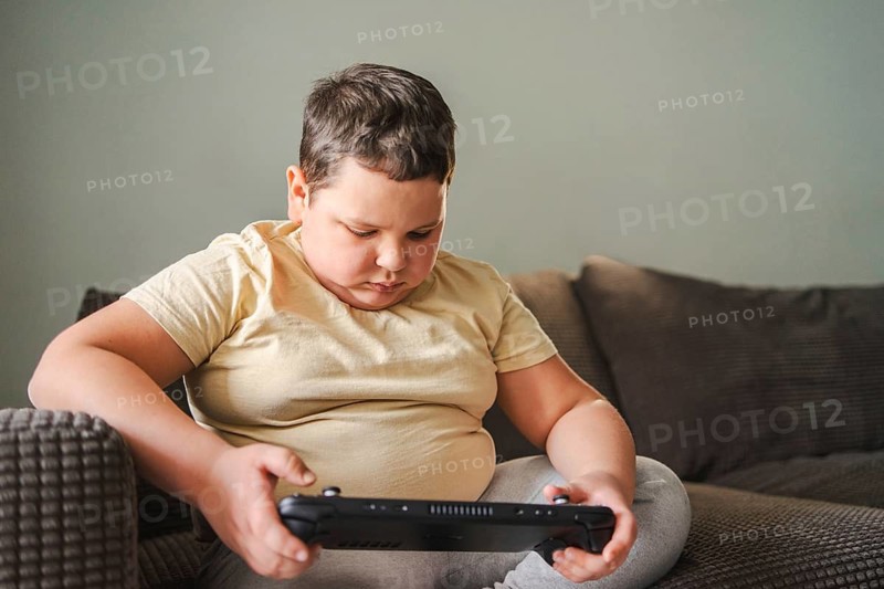 Young boy focused on playing a hand held gaming console while sitting comfortably on a cushioned sofa. The scene captures the essence of leisure and entertainment indoors