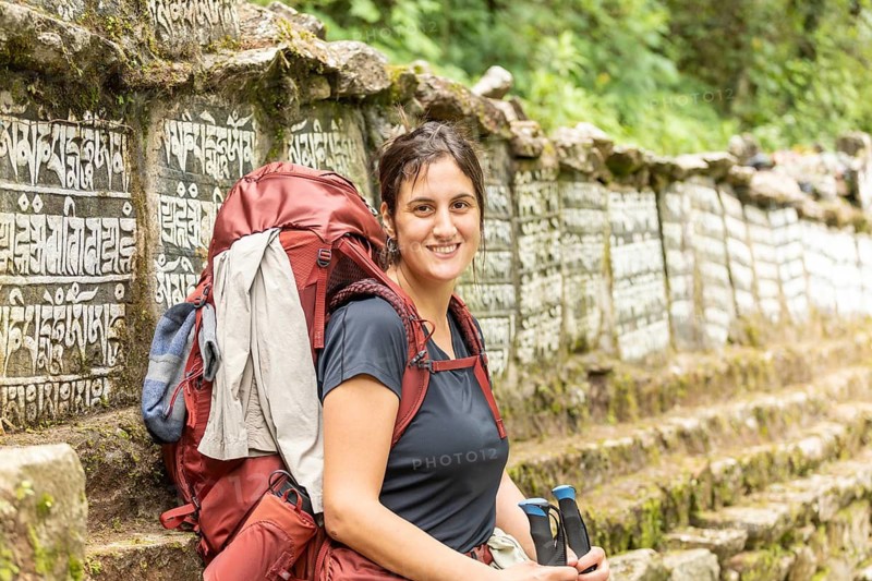 Smiling woman with a backpack trekking along a lush trail near Everest, surrounded by stone walls with carvings, capturing the spirit of adventure and nature's tranquility