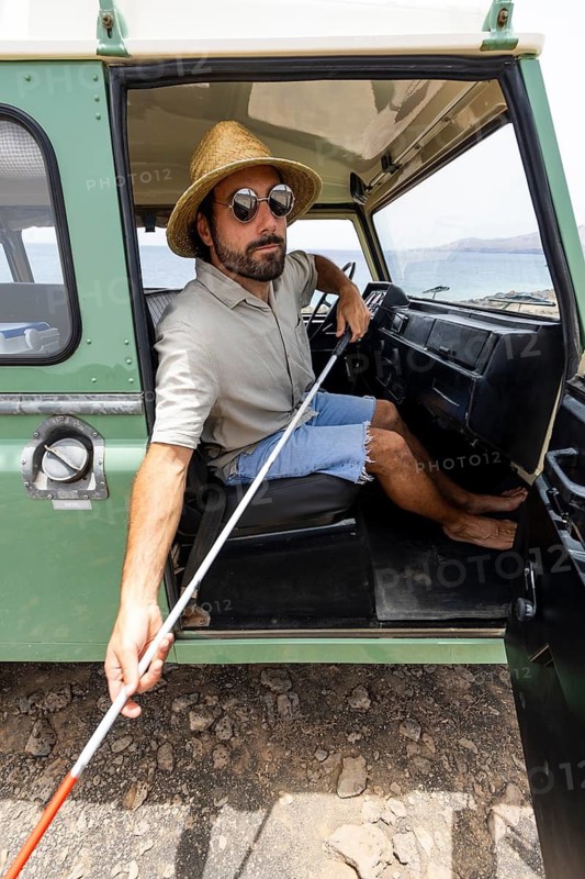 Blind man sitting in a motorhome, wearing sunglasses and a straw hat, holding a cane. He embraces the adventurous spirit and freedom of travel