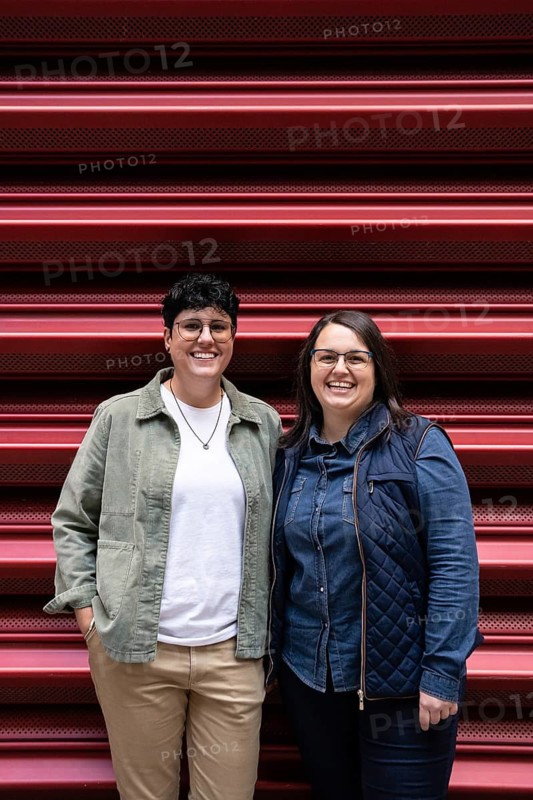 A joyful lesbian couple poses in casual attire against a red backdrop, showcasing their happiness and love. Both women are wearing glasses and smiling warmly