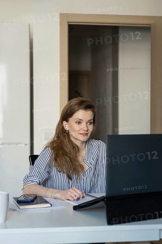 A woman in a striped shirt works on a laptop at home, engaging in a virtual meeting. She appears focused and attentive, using earbuds and taking notes with a notebook nearby