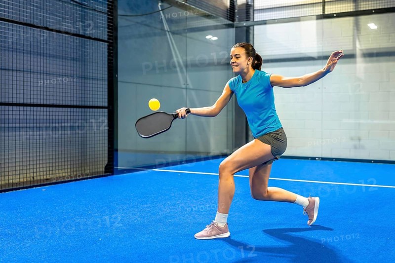 Woman playing pickleball, striking a yellow ball with a paddle, demonstrating agility and focus on a vibrant blue indoor court, embodying sport and healthy lifestyle