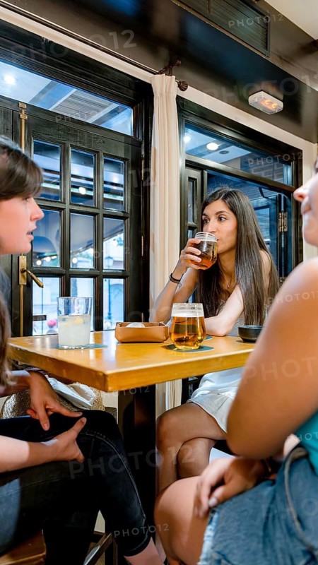 Three friends gather around a wooden table in a cozy restaurant, enjoying refreshing drinks and engaging conversation. The bright atmosphere adds warmth to their evening out