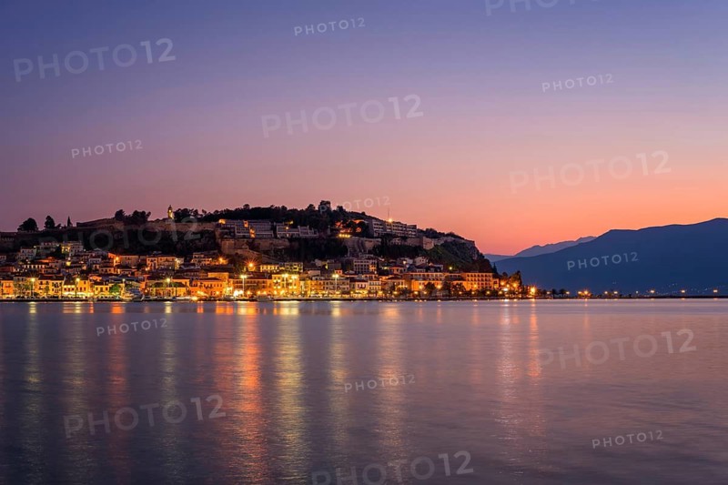 A stunning view of Nafplion, Greece, at sunset with warm city lights reflecting on the calm water. The historic town, nestled on a hillside, creates a picturesque Mediterranean atmosphere