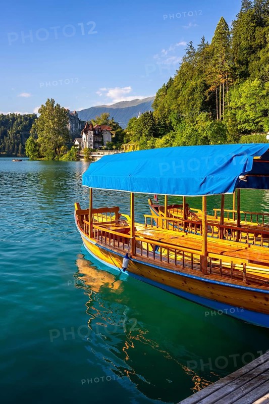 Traditional wooden boat with blue canopy docked on lake bled, with bled castle and forested hillside in the background under a clear blue sky