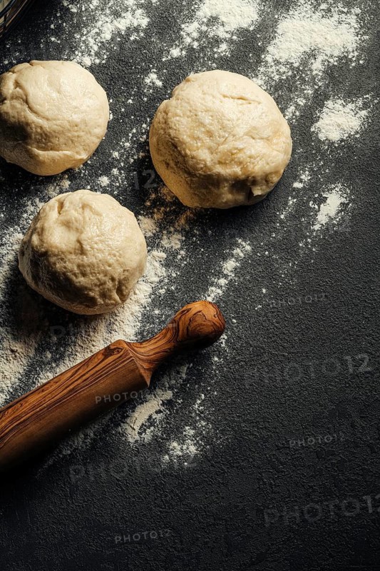 Three dough balls rest on a dark, floured surface, accompanied by a wooden rolling pin, ready for baking. The scene captures the essence of homemade cooking