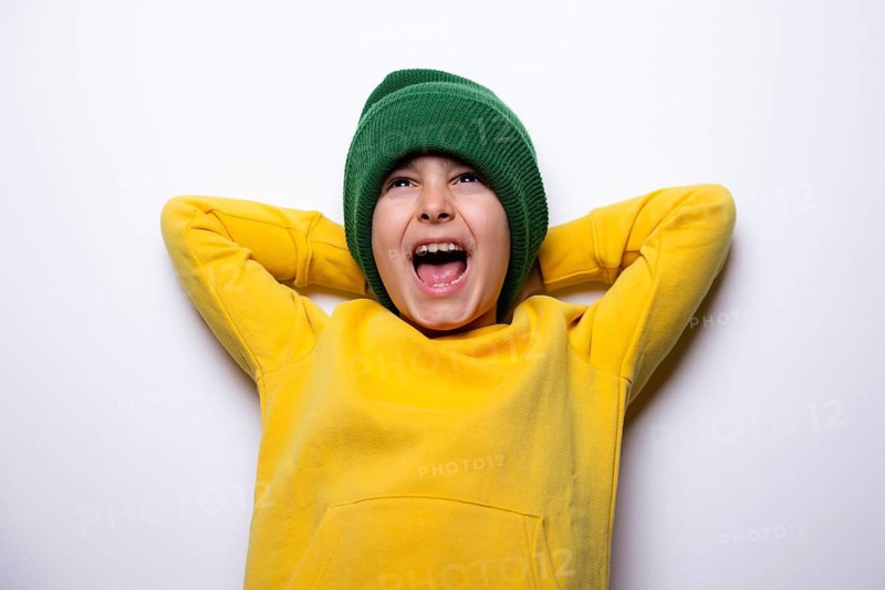 A cheerful child dressed in a vibrant yellow hoodie and a green beanie, arms resting behind head, expressing joy and excitement against a white background