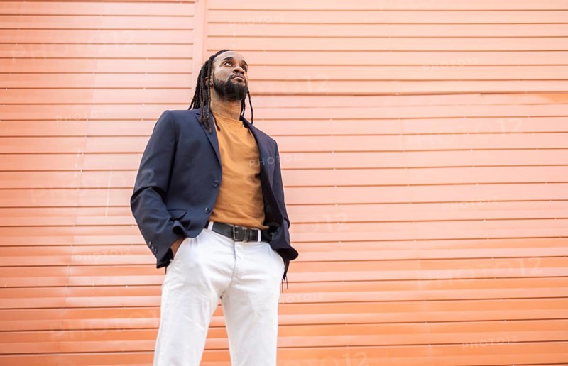 A cuban business man in a dark blazer and orange shirt stands confidently against a textured orange wall. The image captures his upward gaze and relaxed pose, exuding self assurance