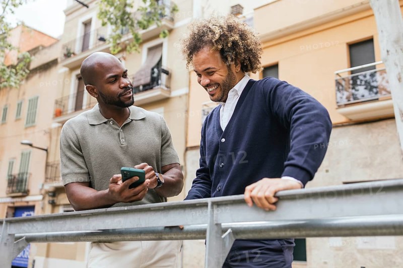 Two men enjoying a break outside a coworking space share a smartphone, engaged in a friendly and relaxed conversation, surrounded by an urban setting with buildings
