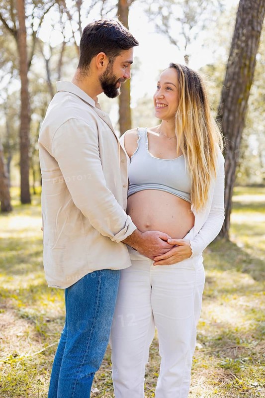 A pregnant woman stands outside with her partner, sharing a joyful moment. They embrace under the trees, expressing love and anticipation for their upcoming addition